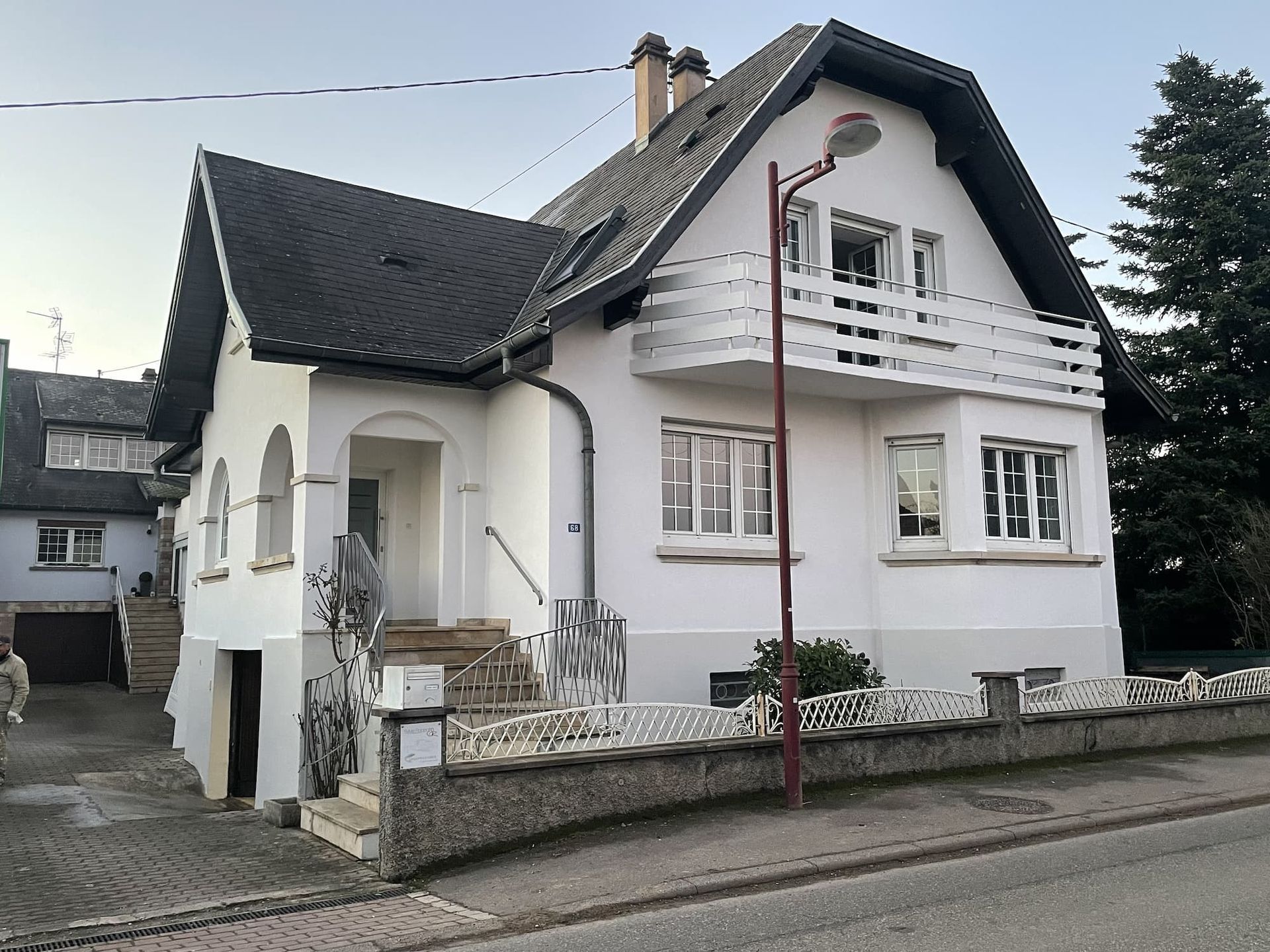 Maison blanche avec toit et balcon sombres, mur en pierre grise, lampadaire.