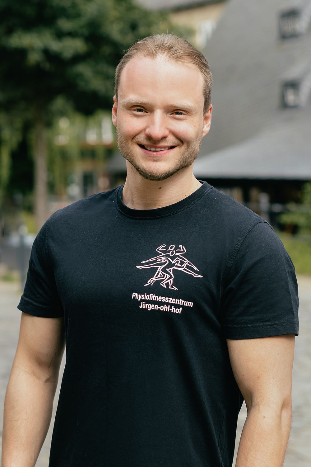 Ein lächelnder Mann in einem schwarzen T-Shirt steht im Freien vor verschwommenen Gebäuden und Grünflächen.