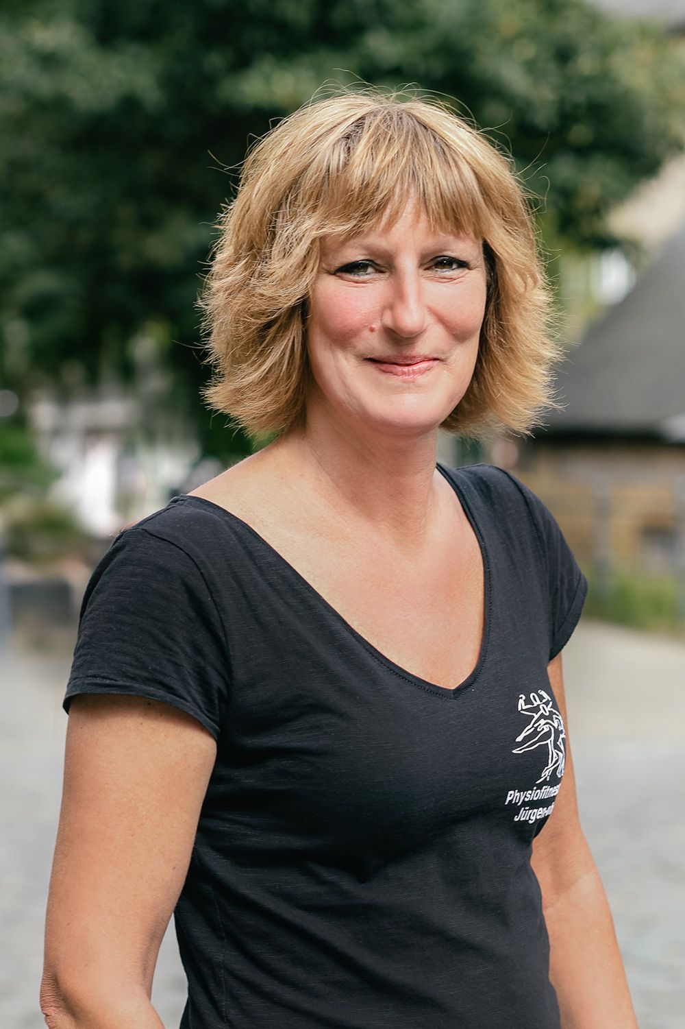 Eine lächelnde Frau in einem schwarzen T-Shirt steht im Freien; im Hintergrund ist verschwommenes Grün zu sehen.
