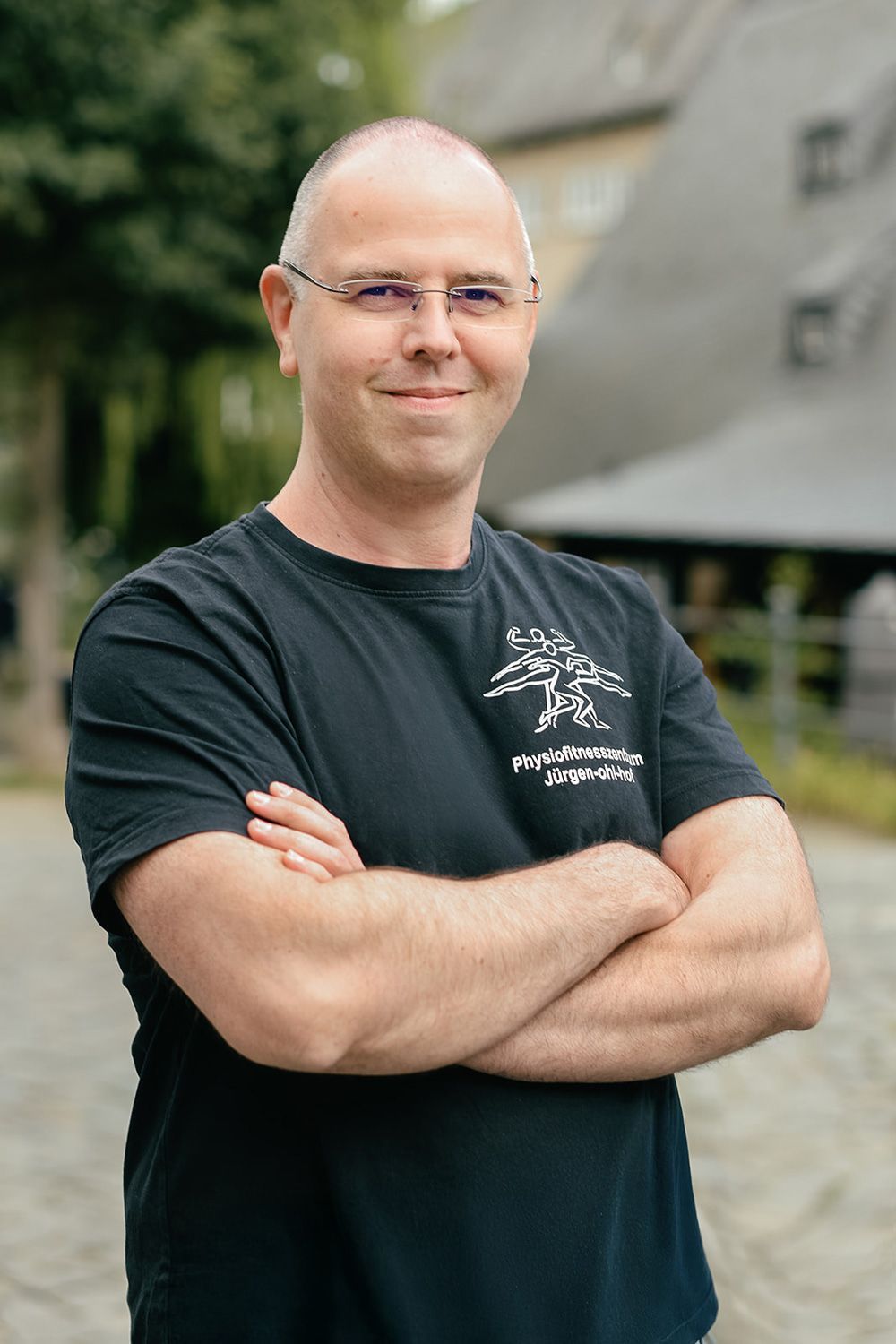 Person mit Brille und verschränkten Armen, bekleidet mit einem schwarzen T-Shirt, steht im Freien