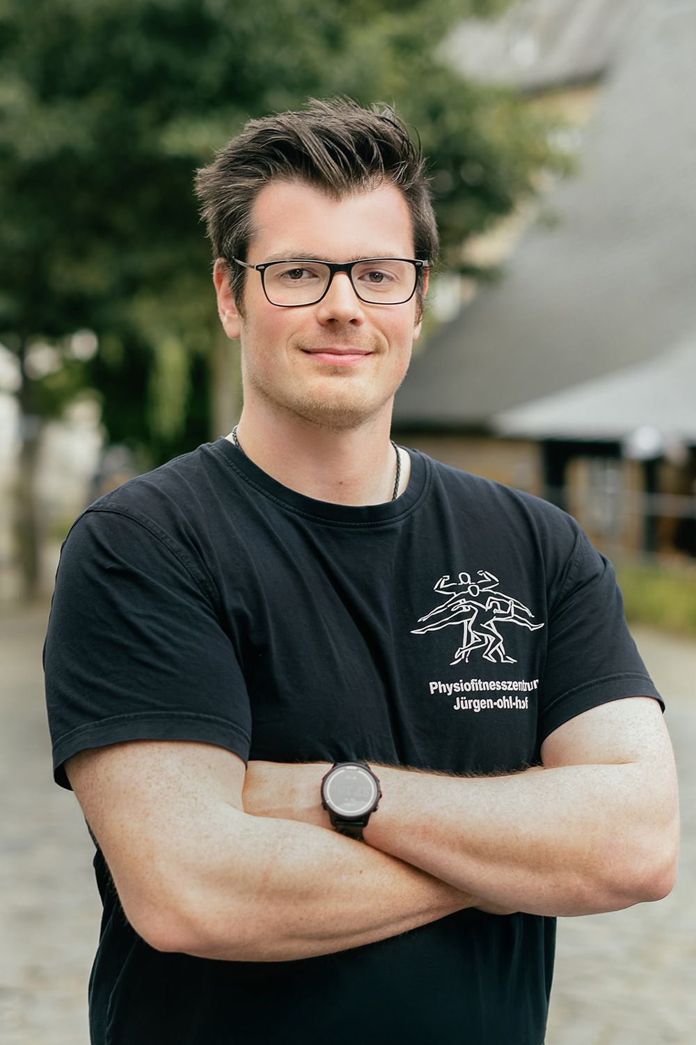 Ein Mann steht mit verschränkten Armen im Freien, trägt ein schwarzes T-Shirt und eine Brille und lächelt in die Kamera.