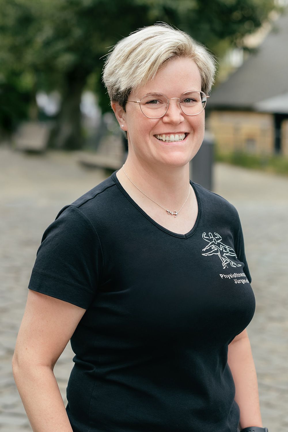 Eine lächelnde Person in einem schwarzen T-Shirt steht im Freien auf einem verschwommenen Pfad;
