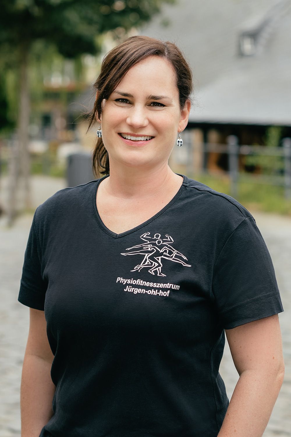 Eine lächelnde Frau in einem schwarzen T-Shirt steht im Freien in der Nähe eines verschwommenen Gebäudes und Grünflächen.