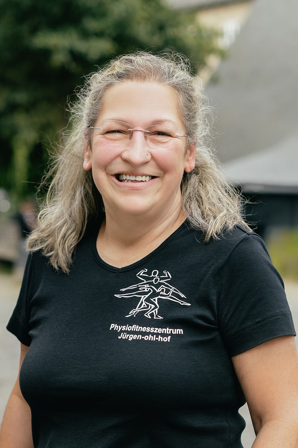 Eine lächelnde Person in einem schwarzen T-Shirt mit weißem Logo steht im Freien vor Grün.