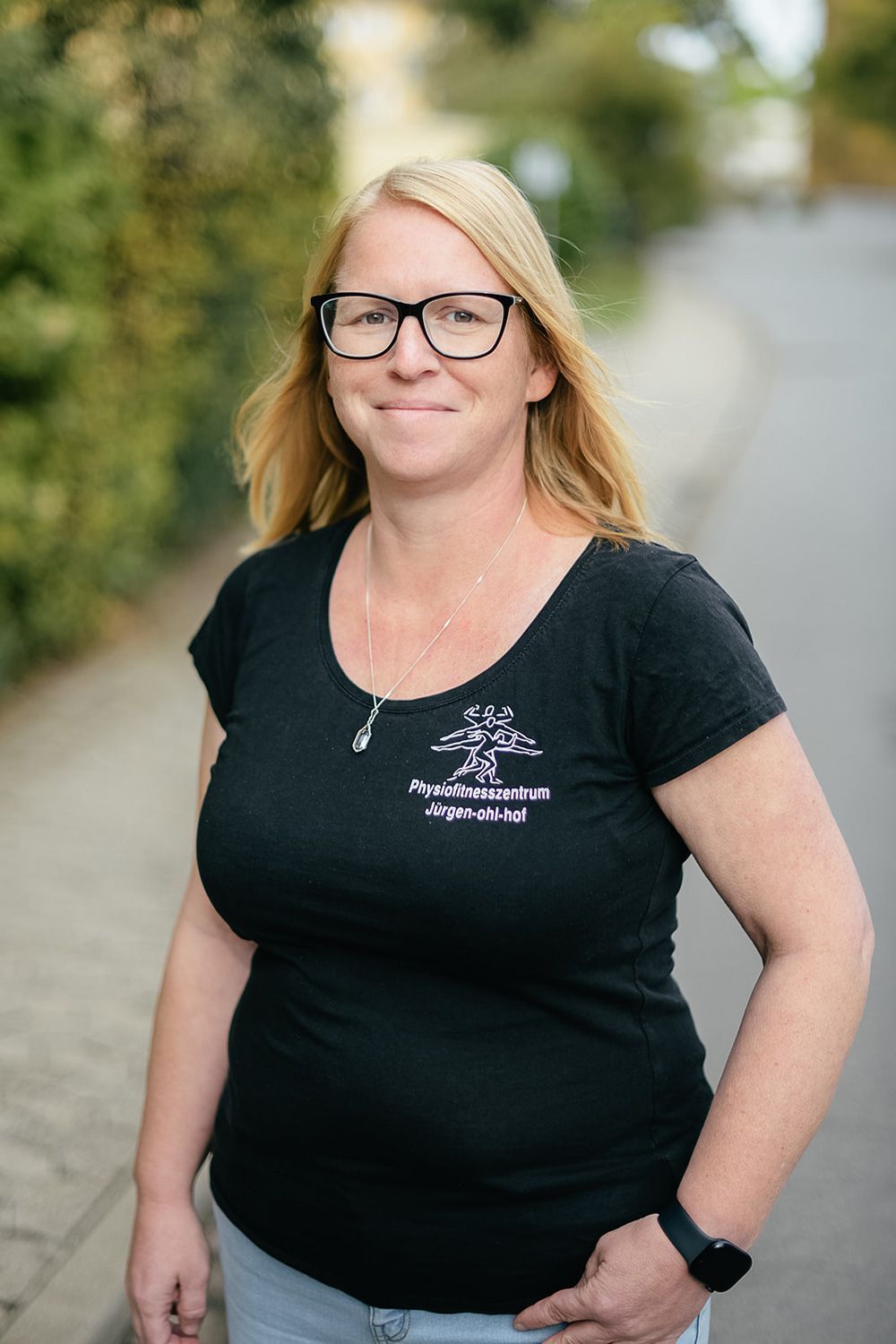 Eine lächelnde Frau mit blonden Haaren und Brille, die ein schwarzes T-Shirt trägt, steht im Freien auf einem Weg.