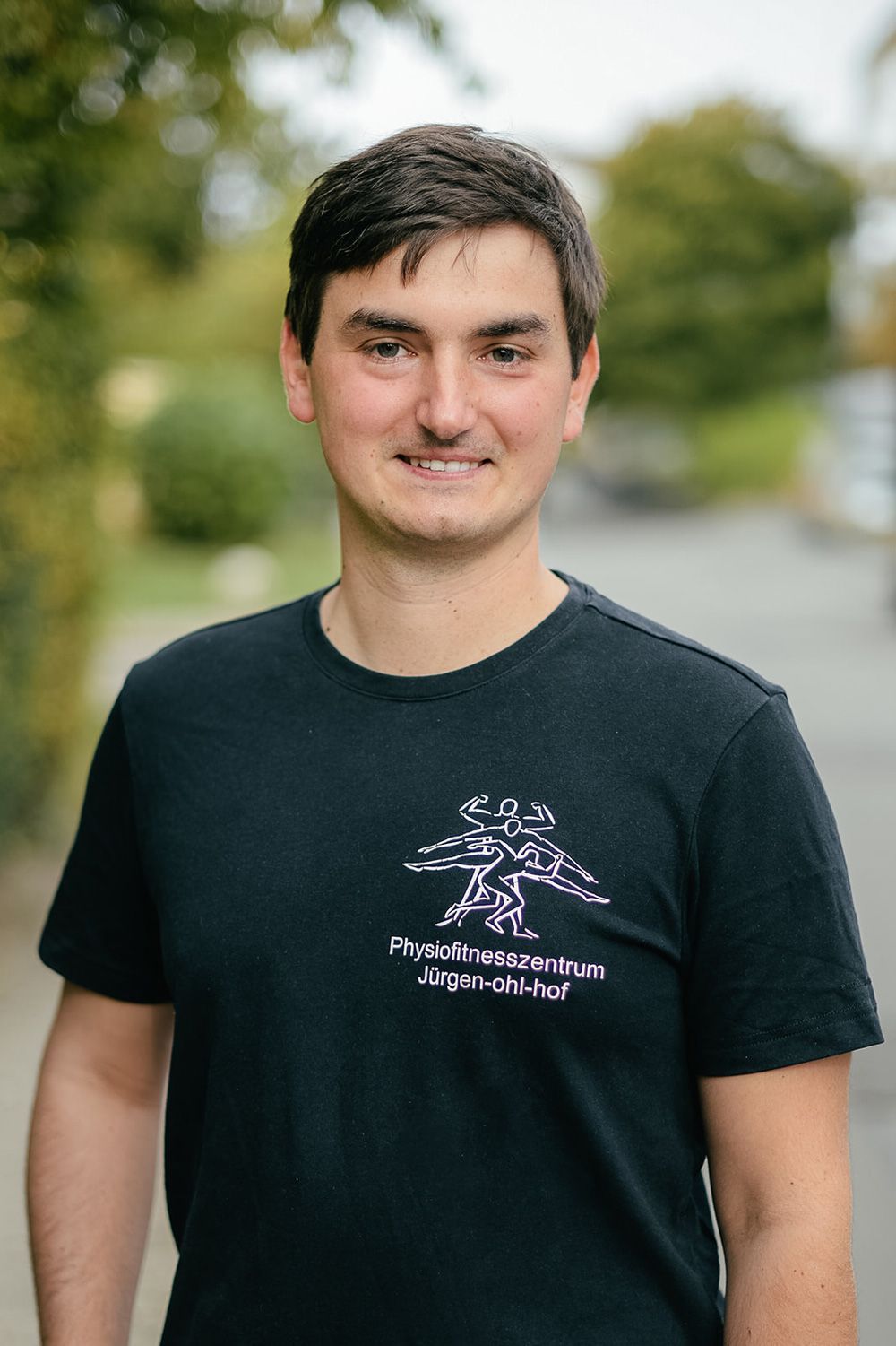 Eine Person im Freien trägt ein schwarzes T-Shirt und steht auf einem Weg vor verschwommenem grünen Hintergrund.