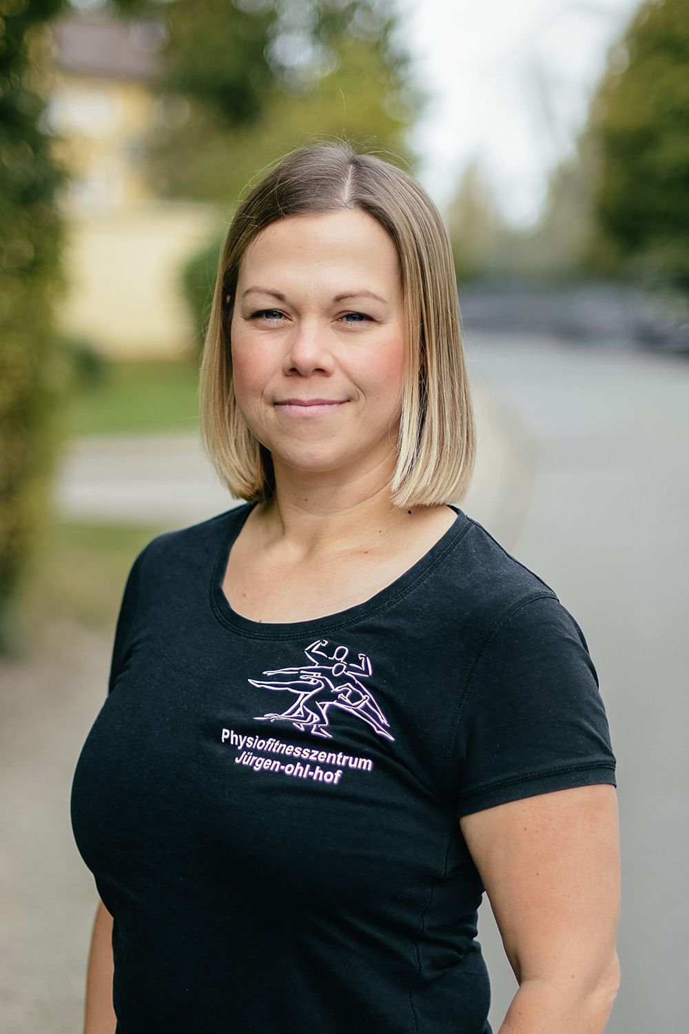 Eine Frau steht im Freien auf einem verschwommenen Pfad. Sie trägt ein schwarzes T-Shirt und hat ein leichtes Lächeln.