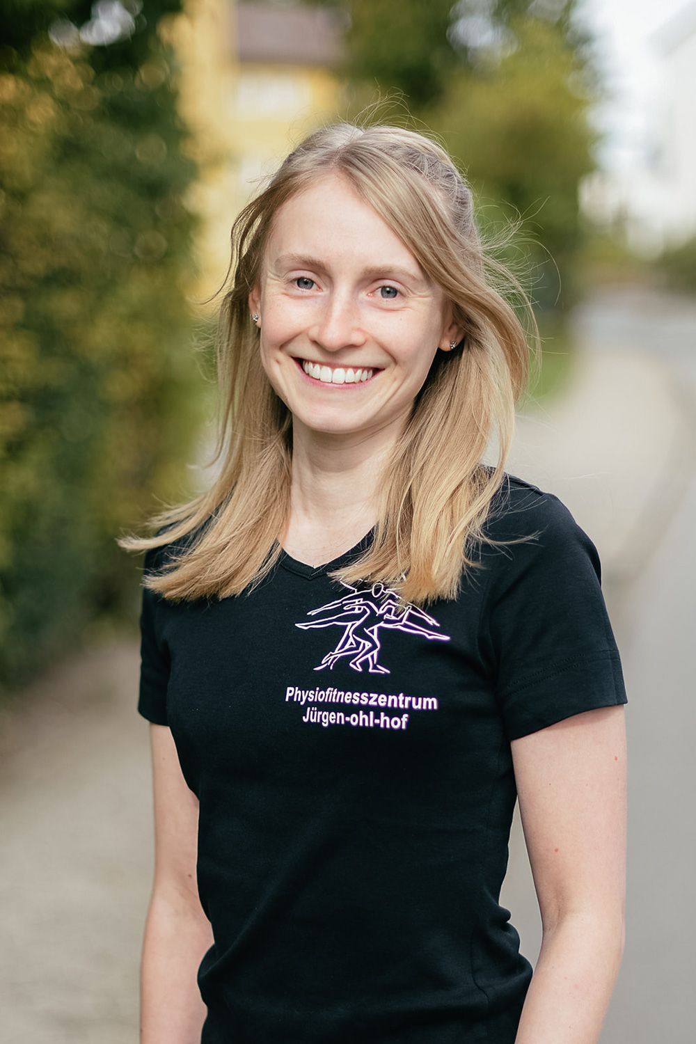 Frau lächelt im Freien; sie trägt ein schwarzes T-Shirt mit pinkfarbenem Logo und Schriftzug.