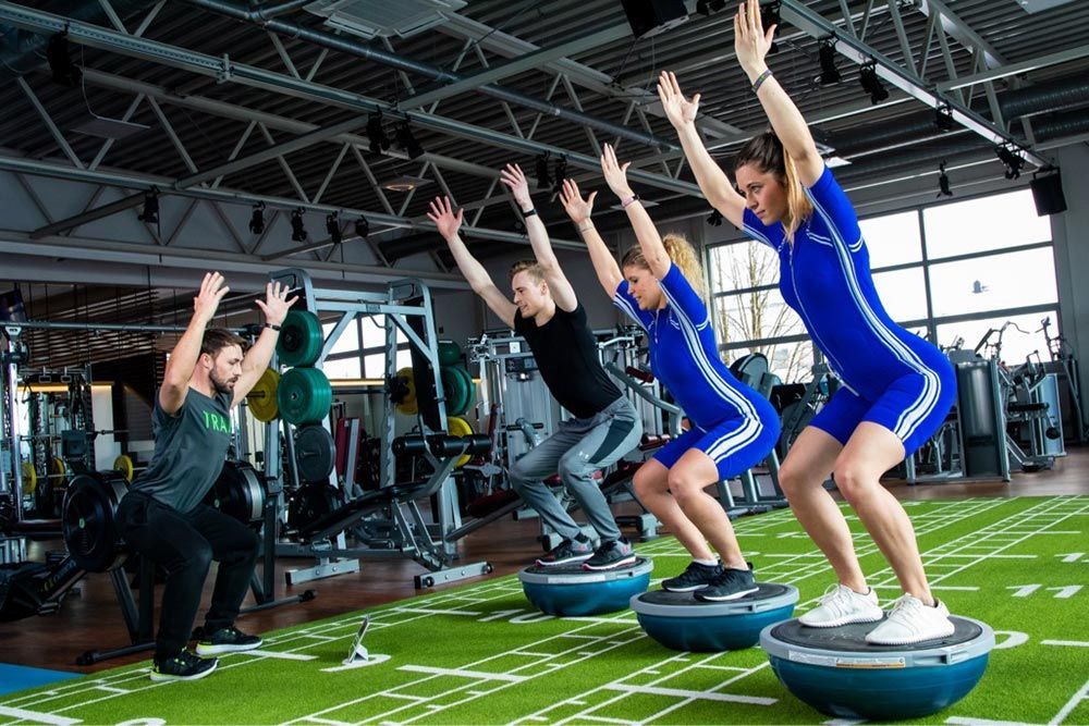 Menschen, die in einem Fitnessstudio Kniebeugensprünge auf Balancetrainern machen, mit erhobenen Armen.