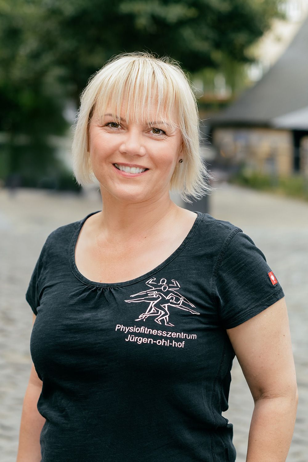 Eine lächelnde Person in einem schwarzen T-Shirt steht im Freien auf einer verschwommenen Straße.