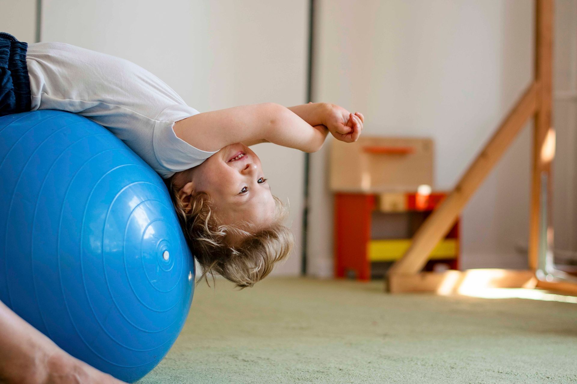 Ein Kind liegt in einem Spielzimmer auf einem blauen Gymnastikball und streckt einen Arm über den Kopf.