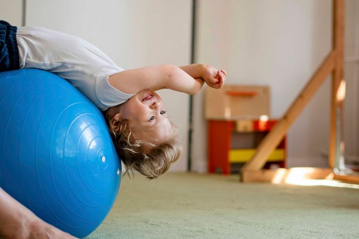 Ein Kind liegt in einem Spielzimmer auf einem blauen Gymnastikball und streckt einen Arm über den Kopf.