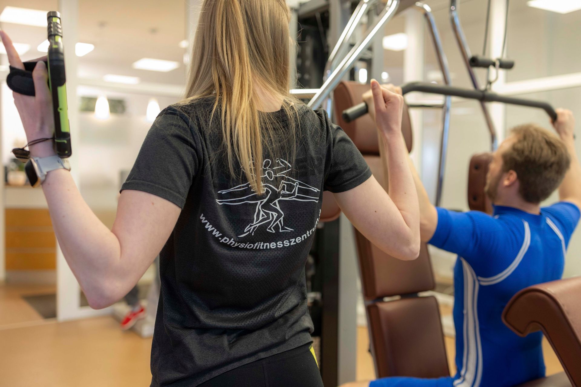 Menschen trainieren an Kraftgeräten in einem Fitnessstudio, im Vordergrund eine Frau und hinter ihr ein Mann in Blau.
