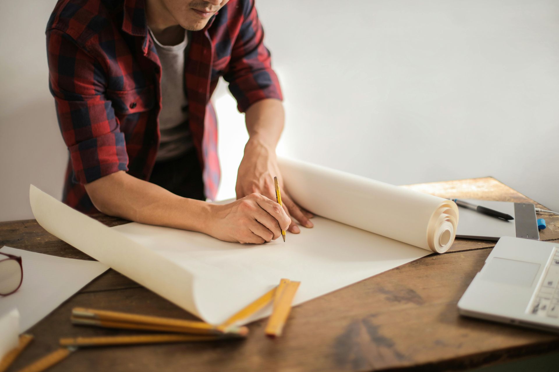 Hombre con camisa a cuadros dibujando en papel, probablemente un arquitecto o diseñador, en un escritorio de madera con herramientas y una computadora portátil.