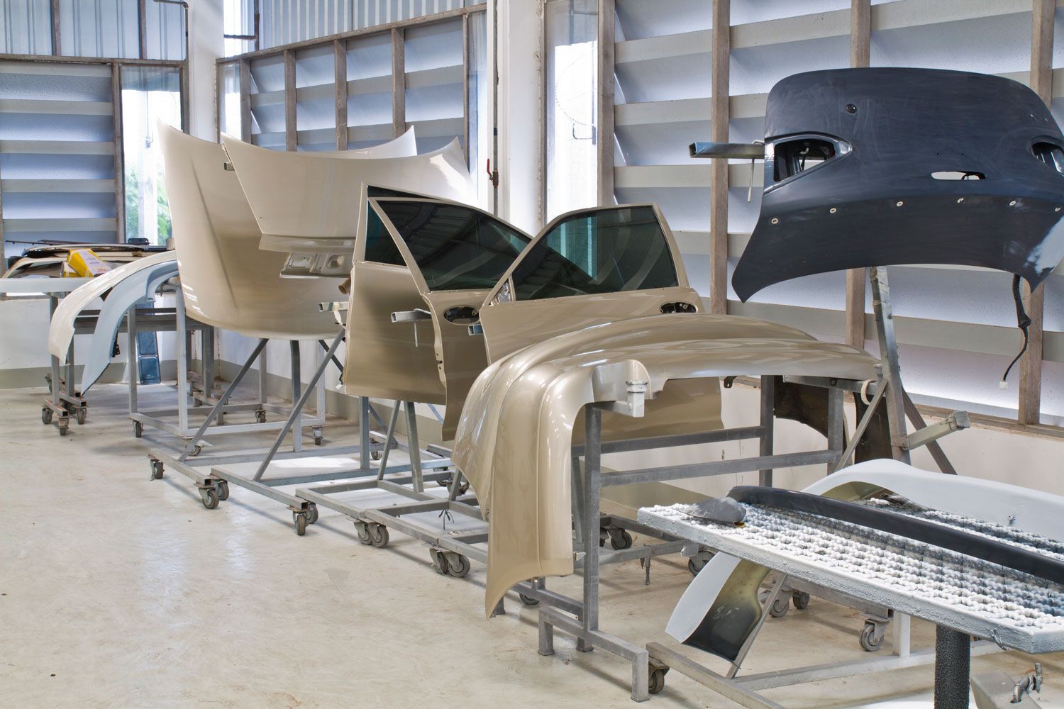 En un taller industrial de carrocería, las piezas de la carrocería del automóvil, incluyendo puertas, parachoques y el capó, reposan sobre soportes metálicos con ruedas.