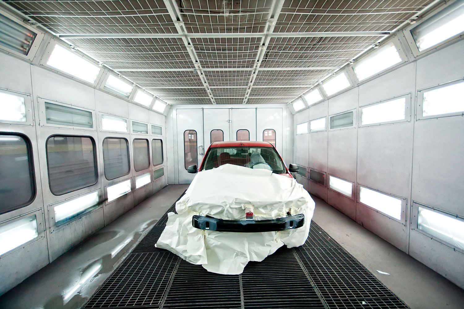 Un coche parcialmente camuflado dentro de una cabina de pintura industrial muy iluminada con suelo de rejilla metálica.