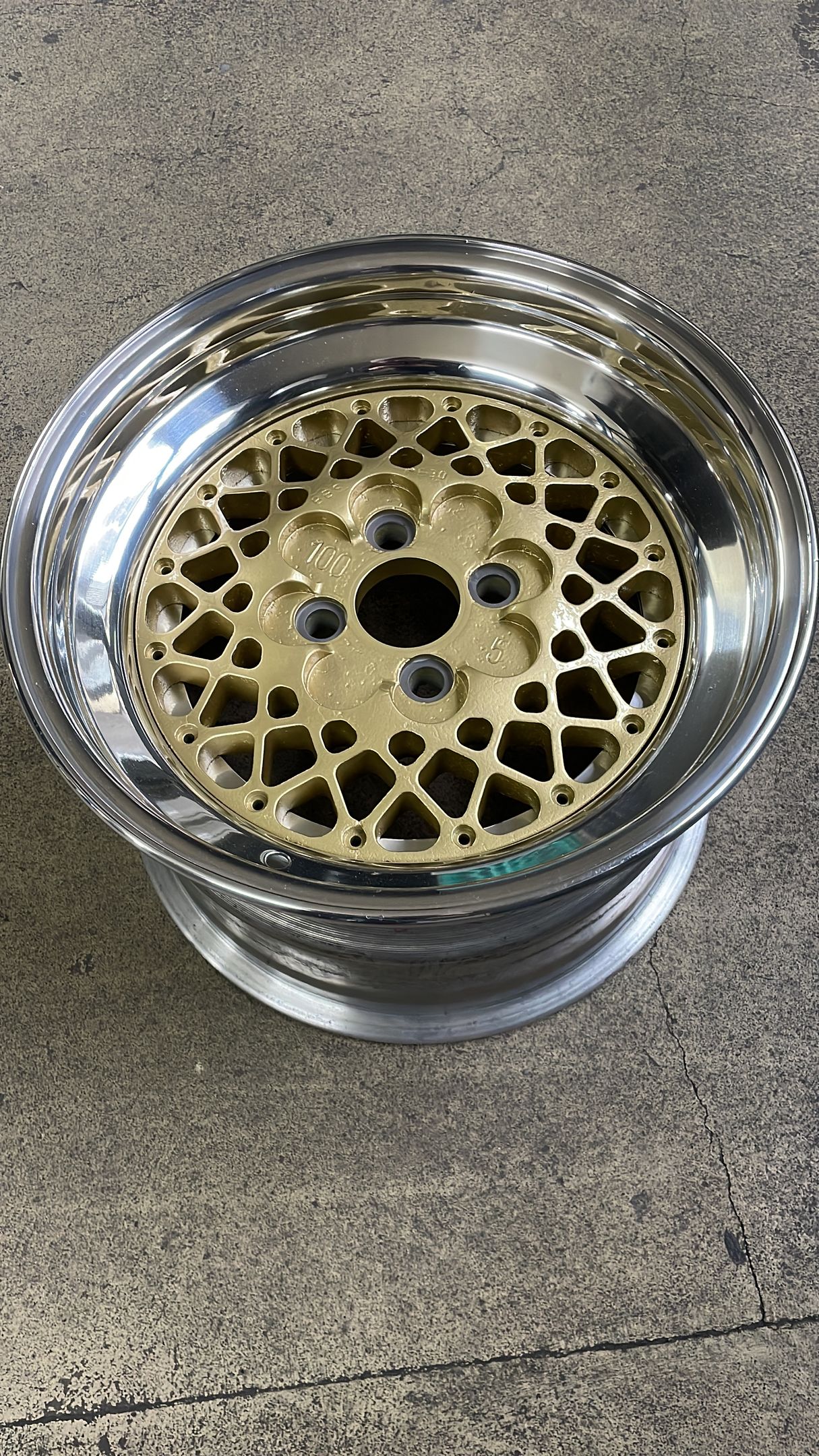 Ein goldenes und verchromtes Autorad mit geometrischem Speichenmuster, das auf einer Betonfläche steht.