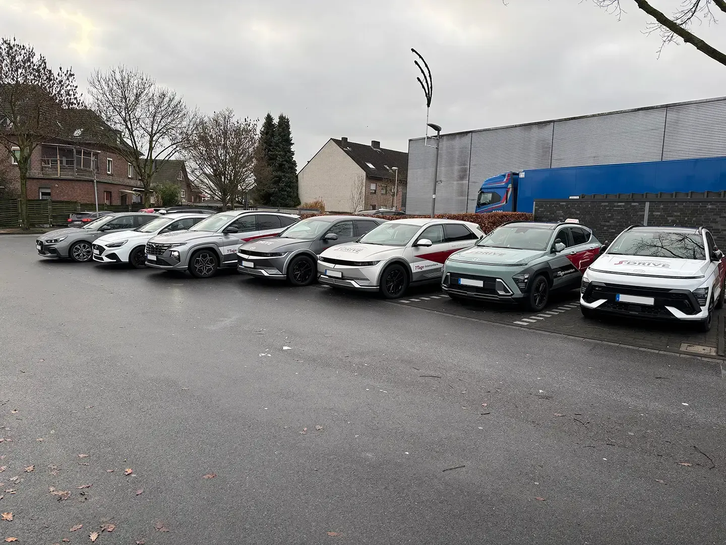 Zwei graue Autos mit „S-DRIVE“-Logos parken vor einem Gebäude. Ein Schild mit der Aufschrift „Führerscheinausbildung in 7 Tagen“ ist zu sehen.