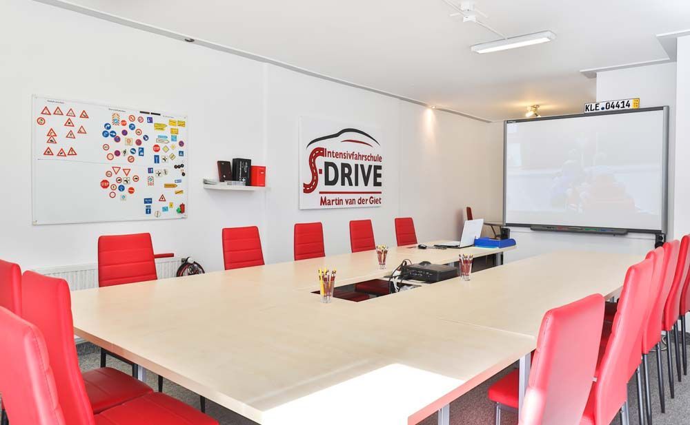 Klassenzimmer mit roten Stühlen, einem langen Tisch, einer Tafel und dem „DRIVE“-Logo an der Wand.