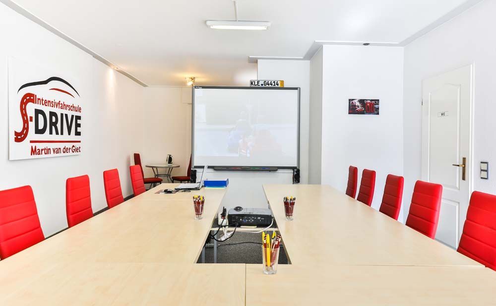 Konferenzraum mit langem Tisch, roten Stühlen und Leinwand. Weiße Wände, Logo der Fahrschule.