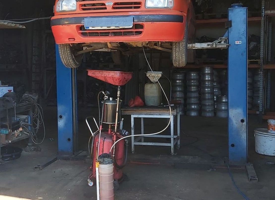 Coche elevado sobre un elevador hidráulico azul en un taller mecánico, con una motocicleta roja debajo y neumáticos apilados en la parte trasera.