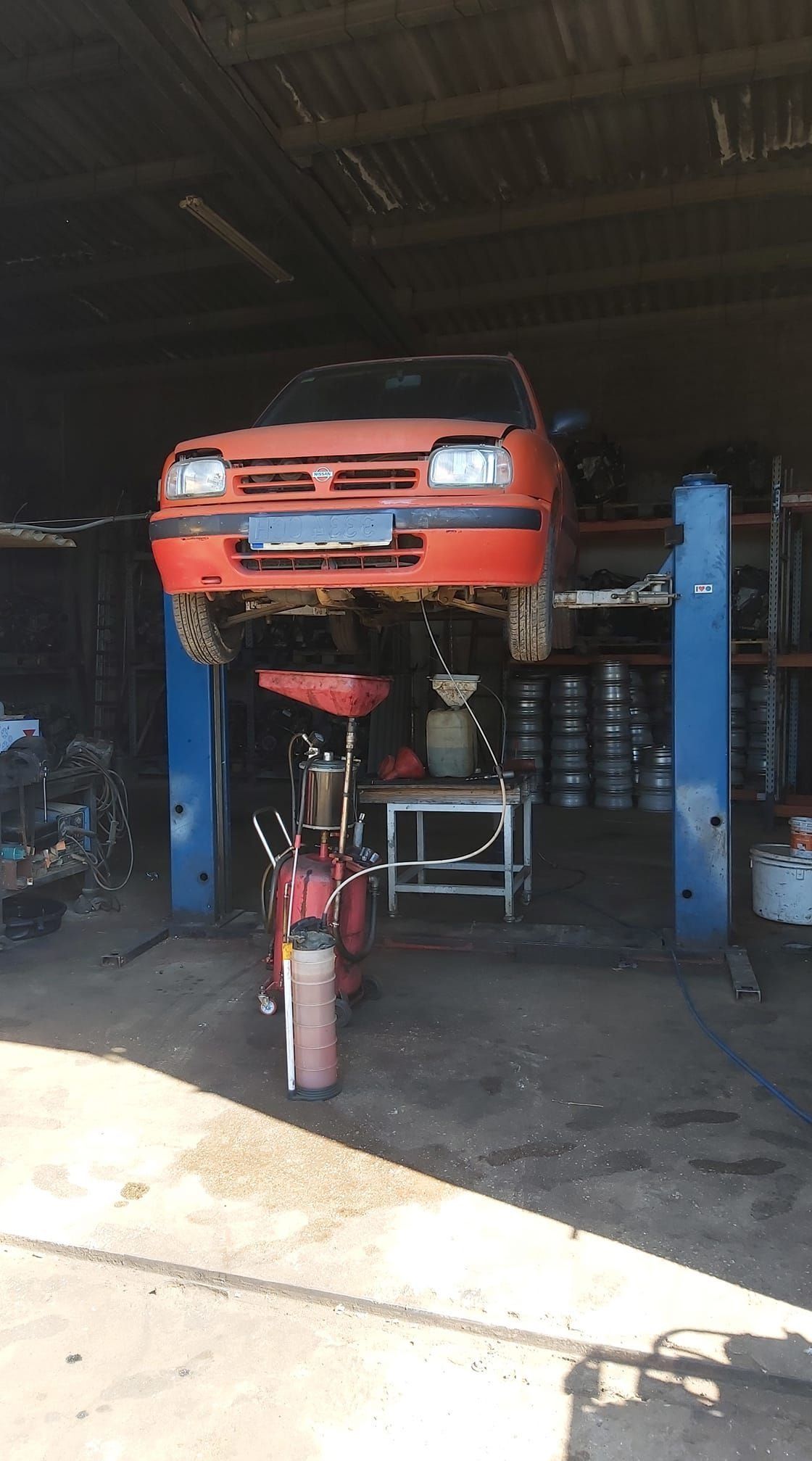Un coche rojo elevado con una grúa hidráulica dentro de un taller mecánico, con herramientas y equipos debajo.