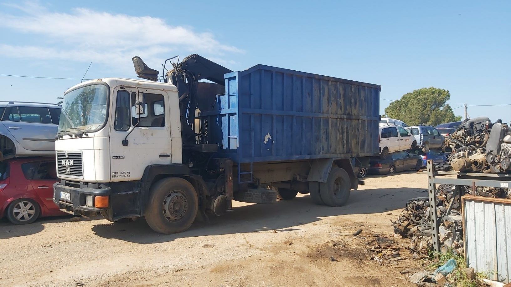 Camión volquete blanco con caja de carga azul estacionado en un desguace en un día soleado.