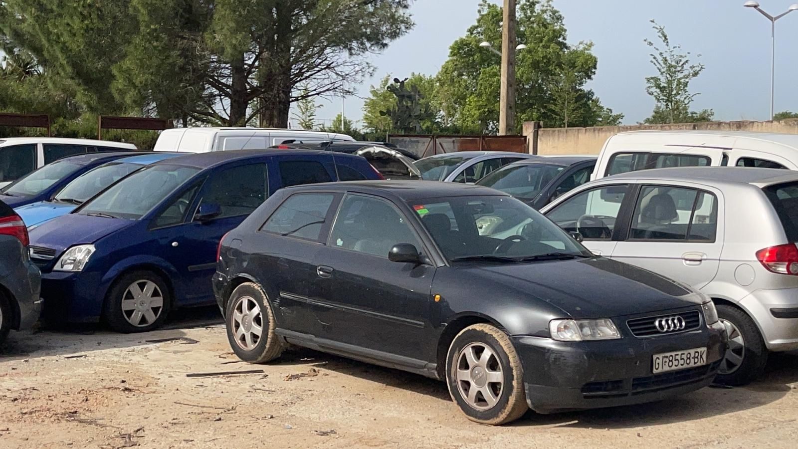 Coches aparcados en un solar polvoriento, entre ellos un sedán gris oscuro en primer plano.