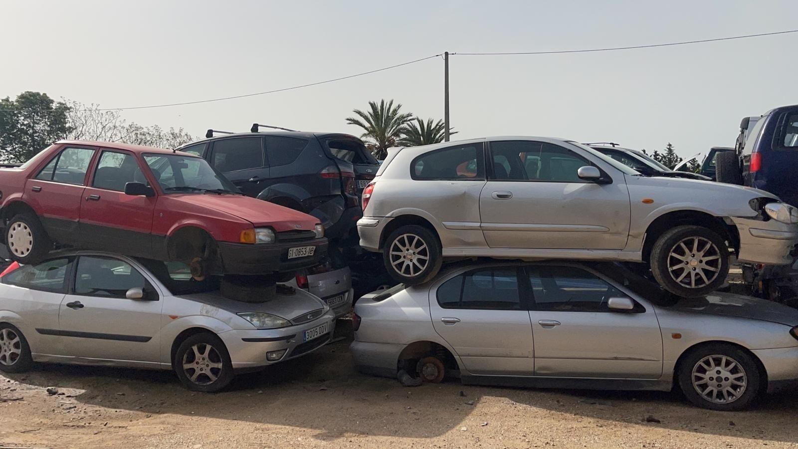 Cuatro coches apilados en un desguace, entre ellos un sedán rojo y un todoterreno beige encima.