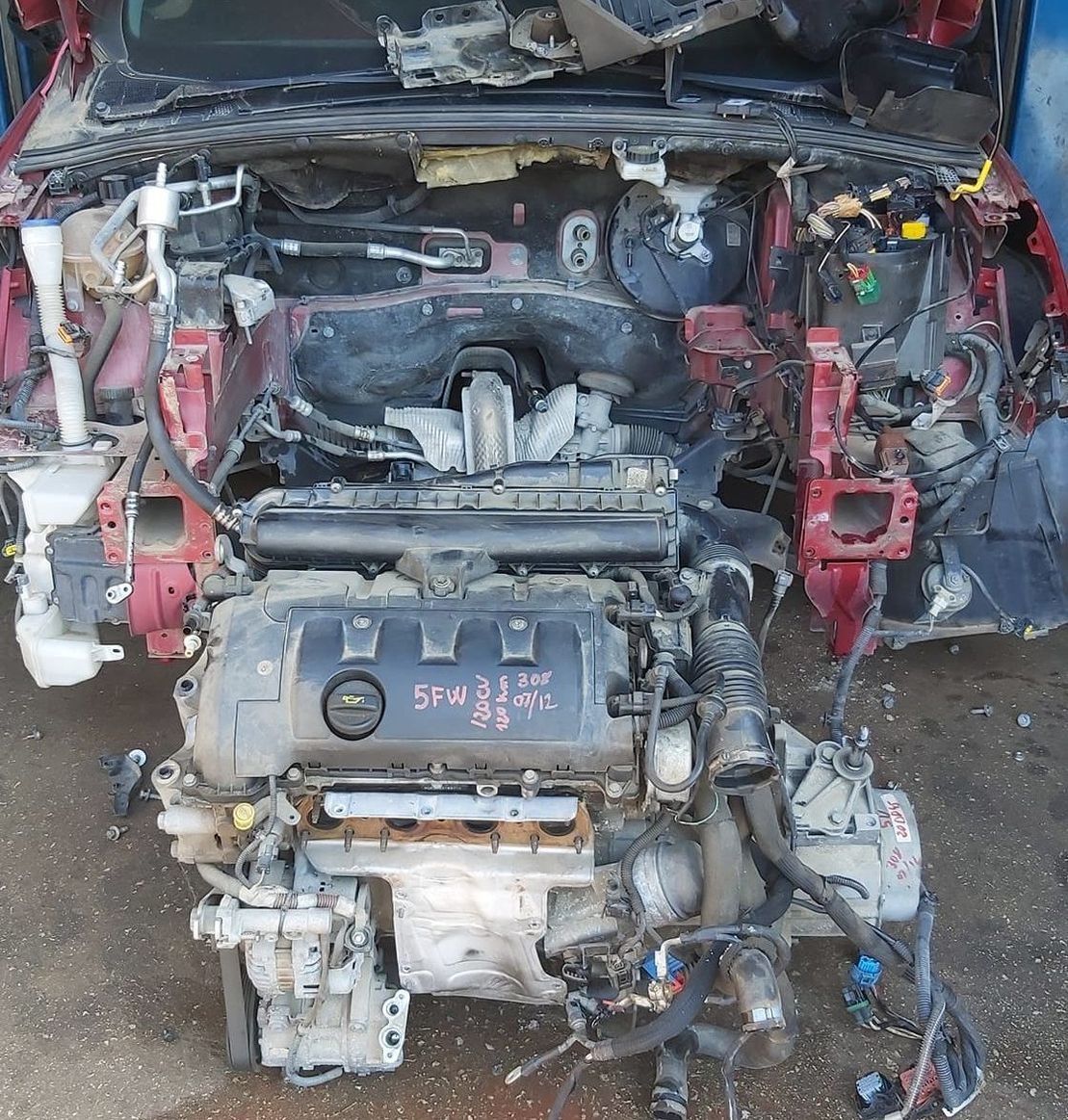 Compartimento del motor del coche desmontado, con piezas expuestas, componentes metálicos sucios y paneles de carrocería rojos bajo el capó.