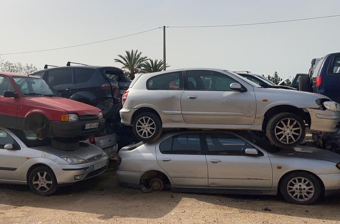 Autos dañados apilados en un desguace, incluyendo un SUV plateado encima de un sedán plateado y un auto rojo detrás.