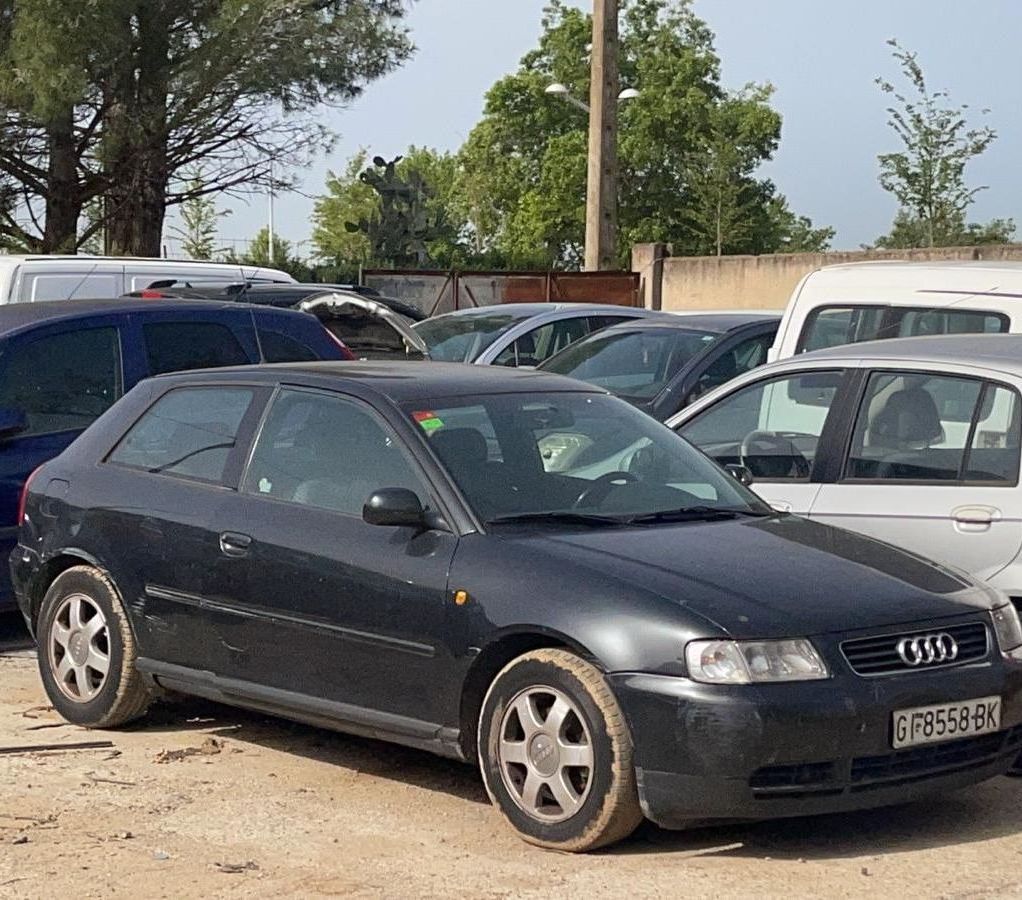 Un sedán Audi negro estacionado en un terreno polvoriento entre otros vehículos, con la parte frontal orientada hacia la cámara.