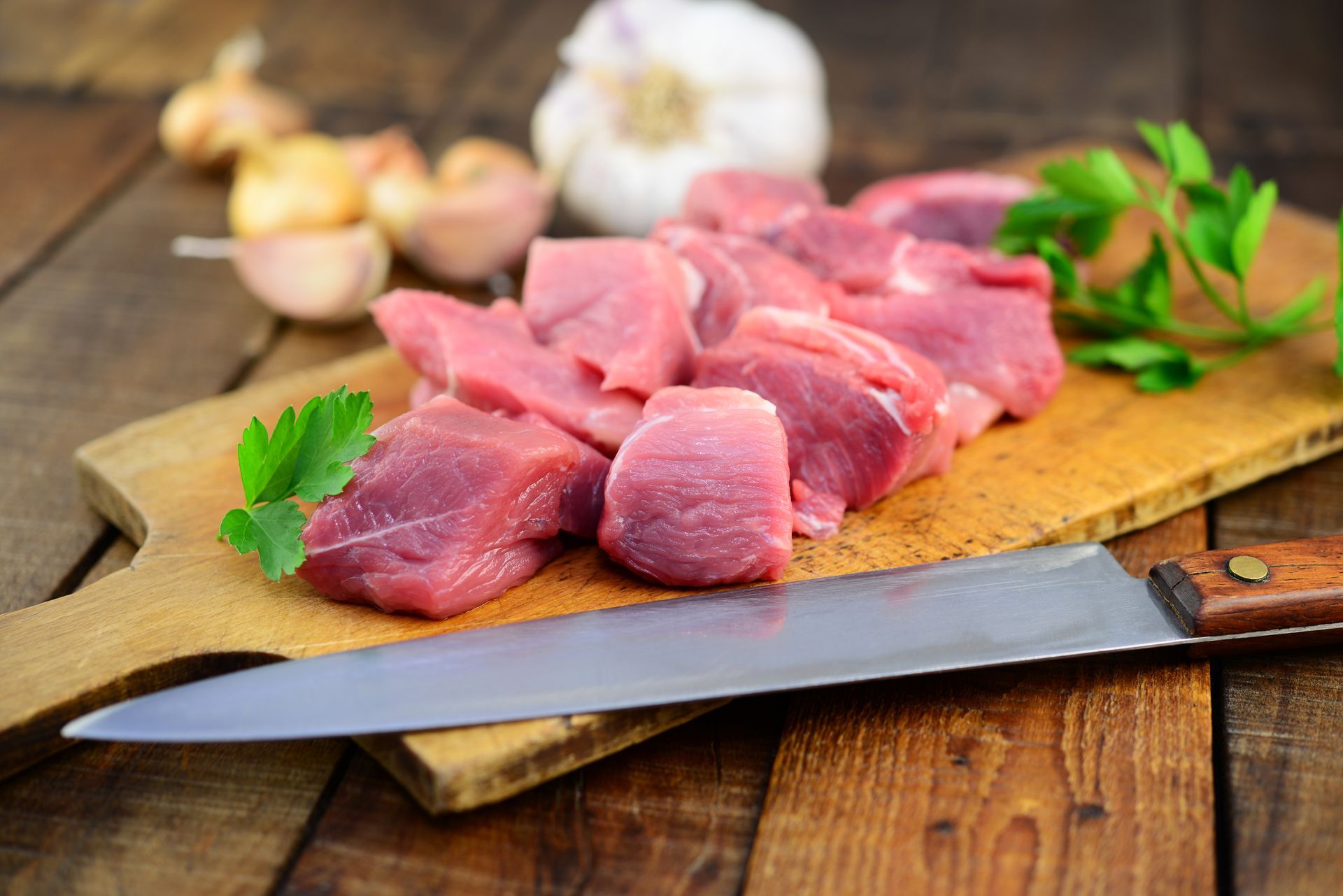 Viande crue coupée en dés sur une planche à découper en bois avec un couteau, de l'ail, des oignons et du persil.