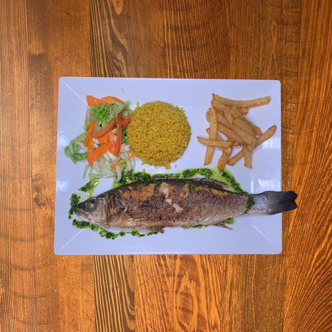 Poisson entier grillé avec riz, frites et salade sur une assiette blanche, table en bois.