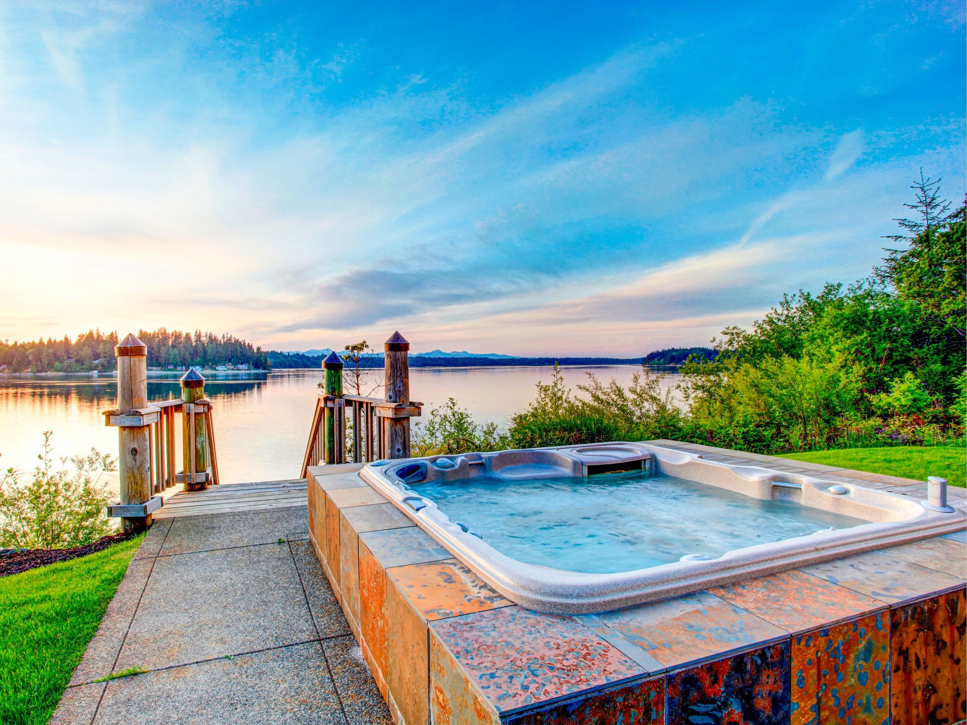 Hot tub overlooking a lake at sunset; wooden dock, blue sky, green grass.