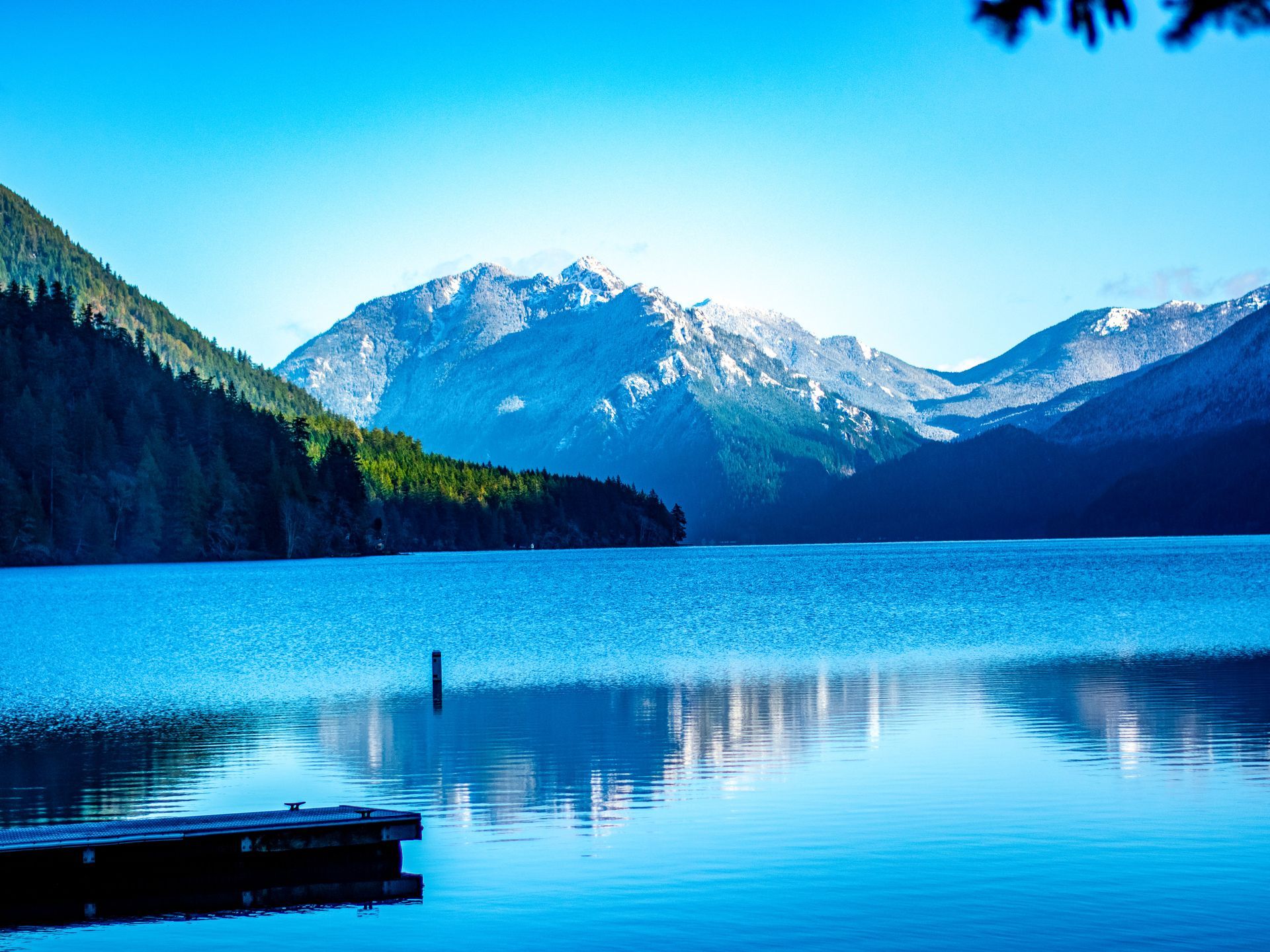 Lake surrounded by mountains, trees, and a wooden dock, blue water under a bright sky.