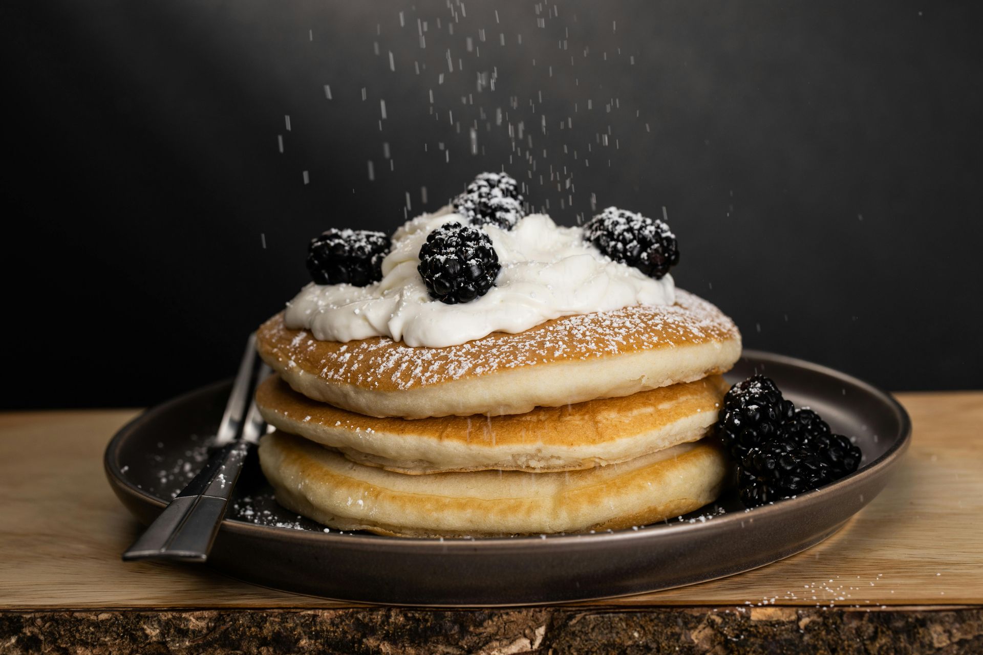 Stack of pancakes with whipped cream, blackberries, and powdered sugar on a brown plate.