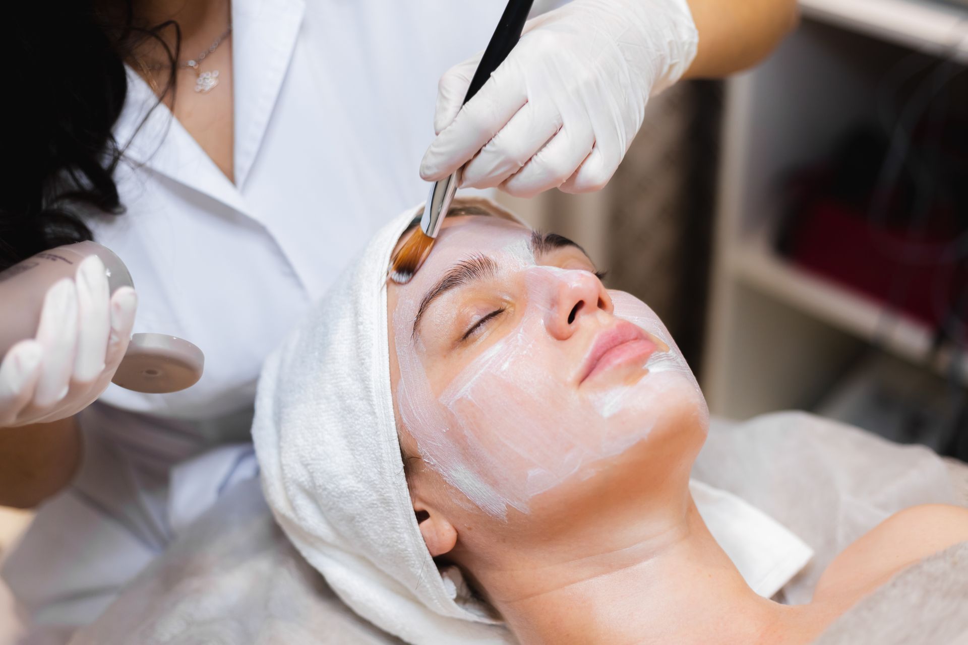 Uma profissional de spa, vestida com uniforme branco e luvas, aplica uma máscara facial cremosa em uma cliente com um pince