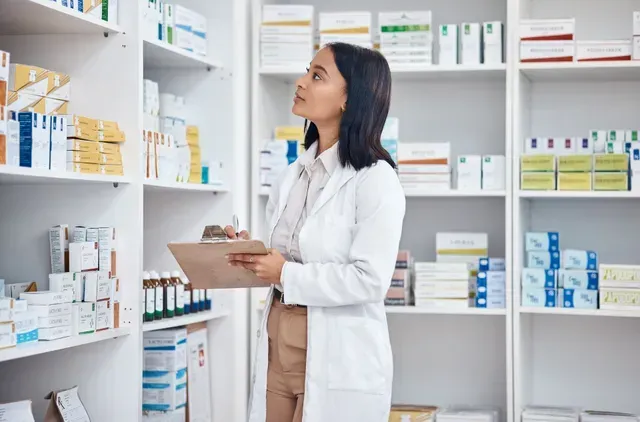 Um farmacêutico de jaleco branco segura uma prancheta e verifica o estoque nas prateleiras de uma farmácia.