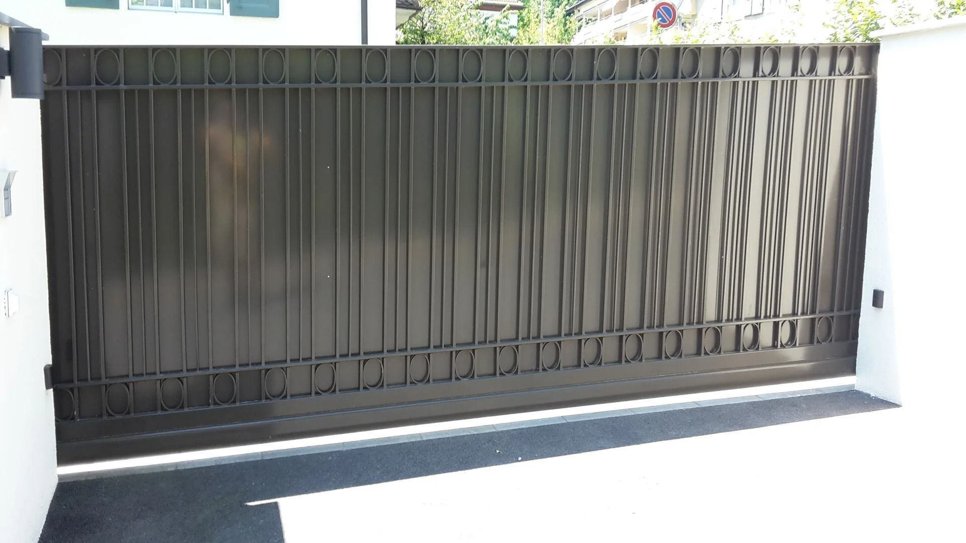 Portail en métal sombre devant un bâtiment blanc, barreaux verticaux et panneaux texturés.