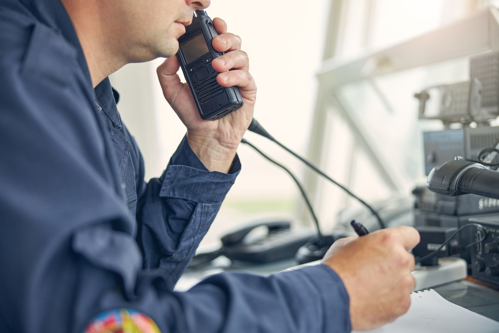 Un homme en uniforme bleu utilise une radio portative, probablement dans une salle de contrôle, en train de communiquer.