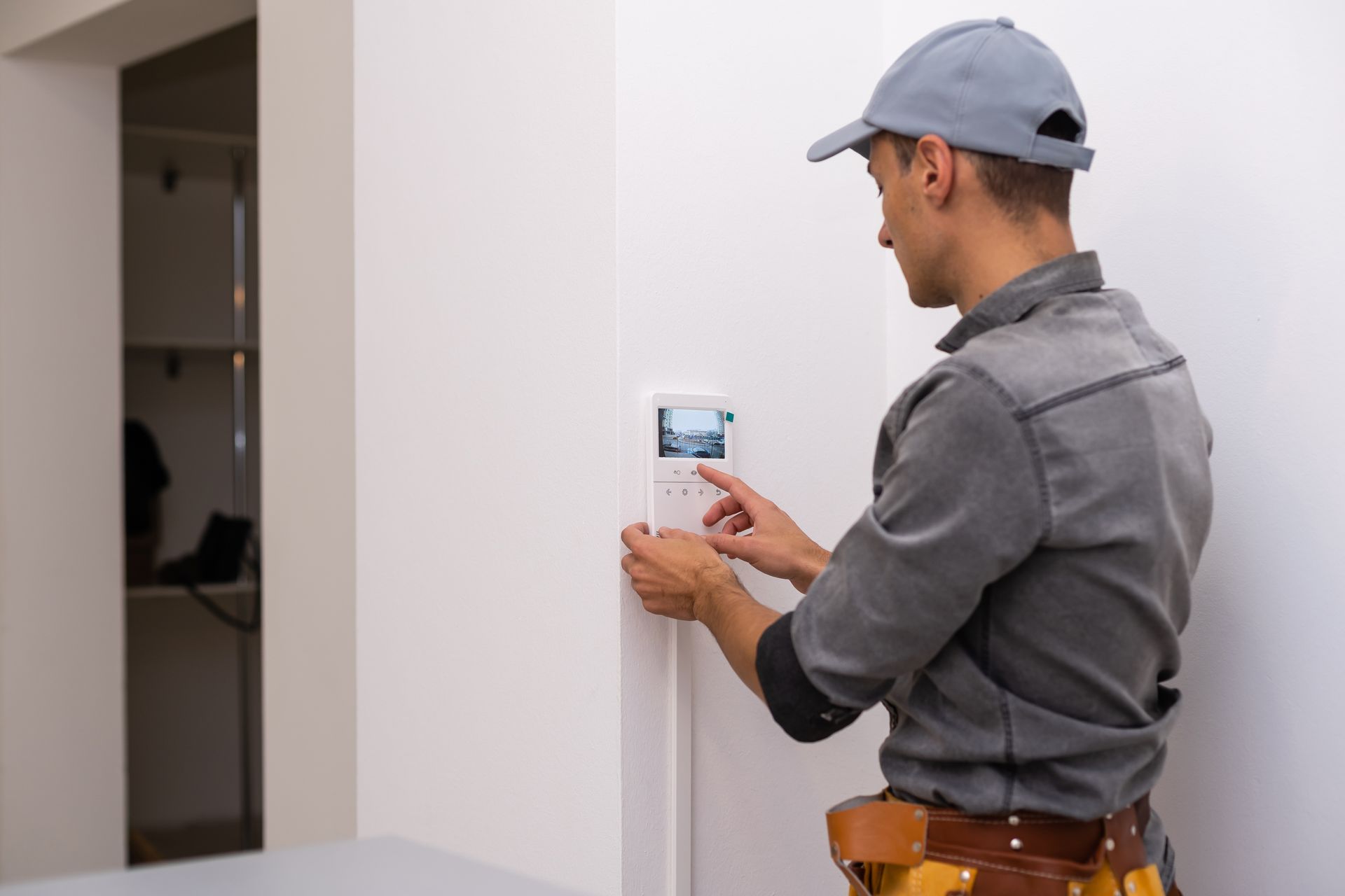 Un homme en uniforme gris avec casquette interagit avec un panneau de contrôle blanc sur un mur.
