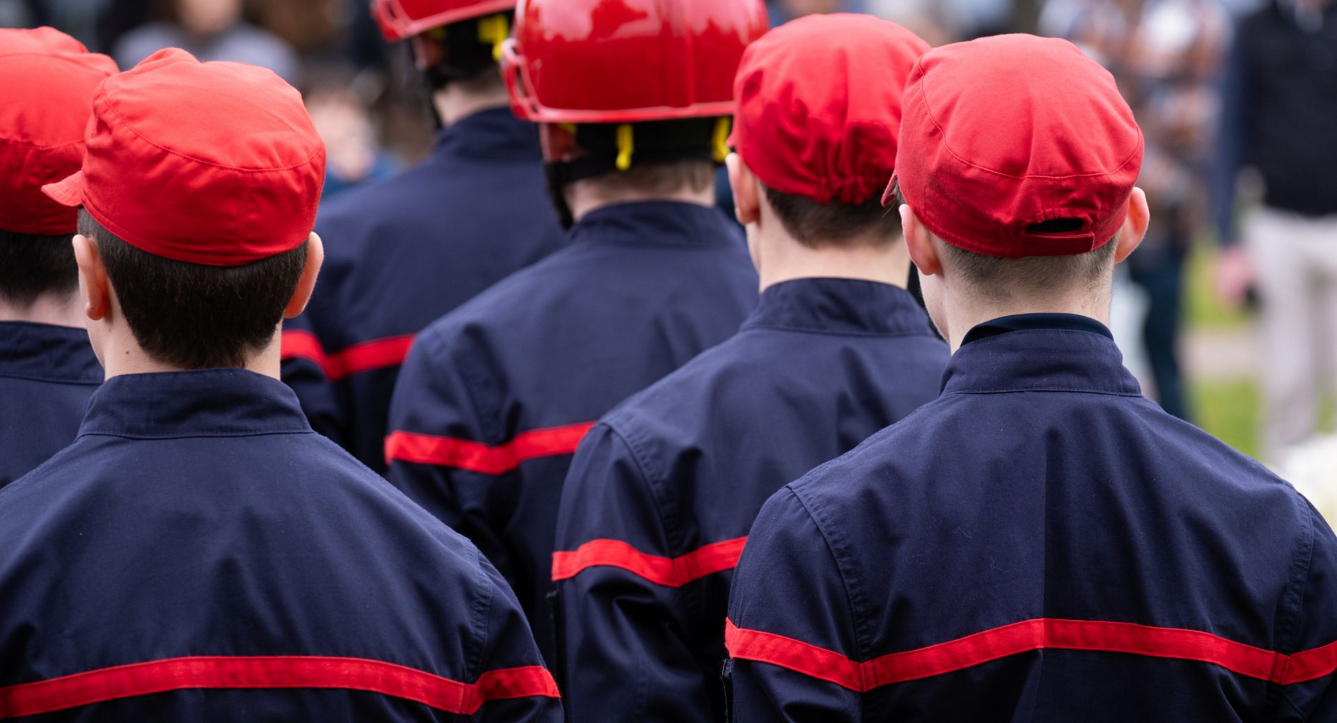 Des pompiers en uniforme bleu foncé et avec une casquette rouge, debout en ligne.