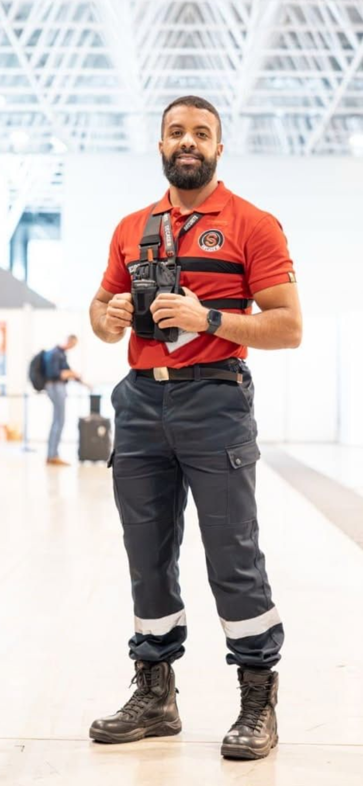 Un homme en uniforme tient un talkie-walkie, probablement dans une salle de contrôle, près d'un bureau avec du matériel.