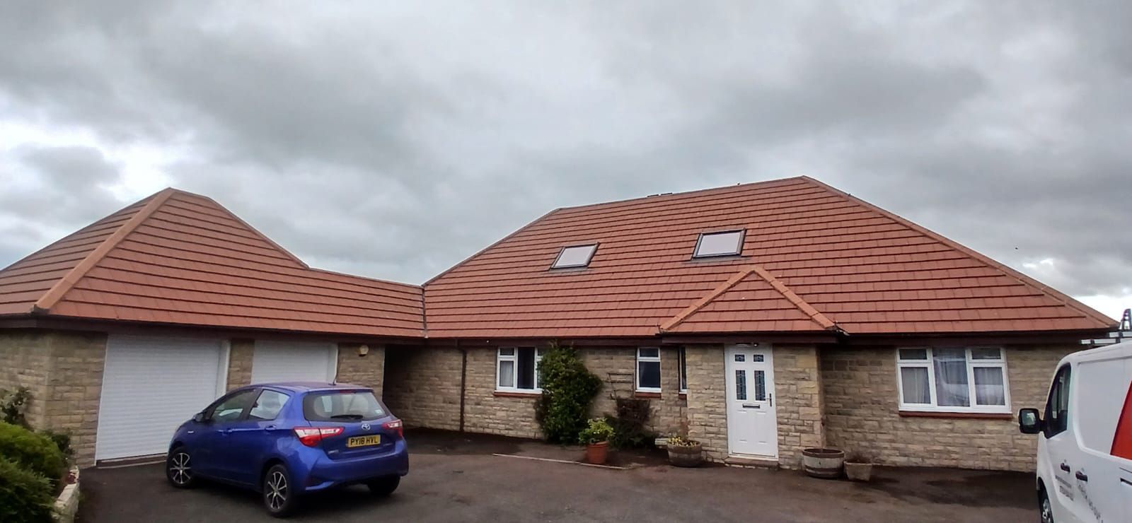 Two single-story houses with red tiled roofs, a blue car parked outside, under a cloudy sky.