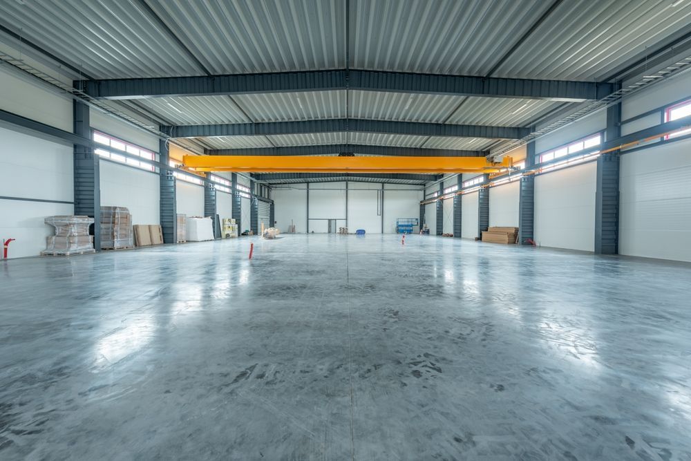Gran almacén industrial vacío con grúa aérea, suelo de hormigón y paredes blancas.