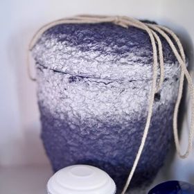 Recipiente texturizado azul escuro e branco com alça de corda, cobrindo parcialmente um objeto branco e um objeto azul.