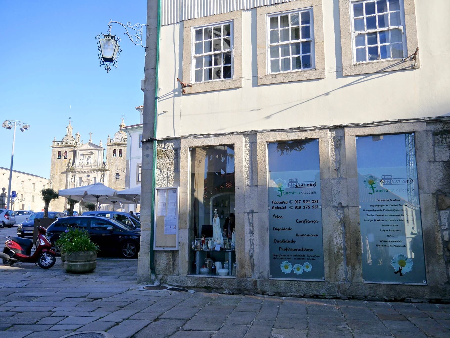 Cena de rua com um edifício de pedra, vitrines, carros estacionados e uma igreja ao fundo.