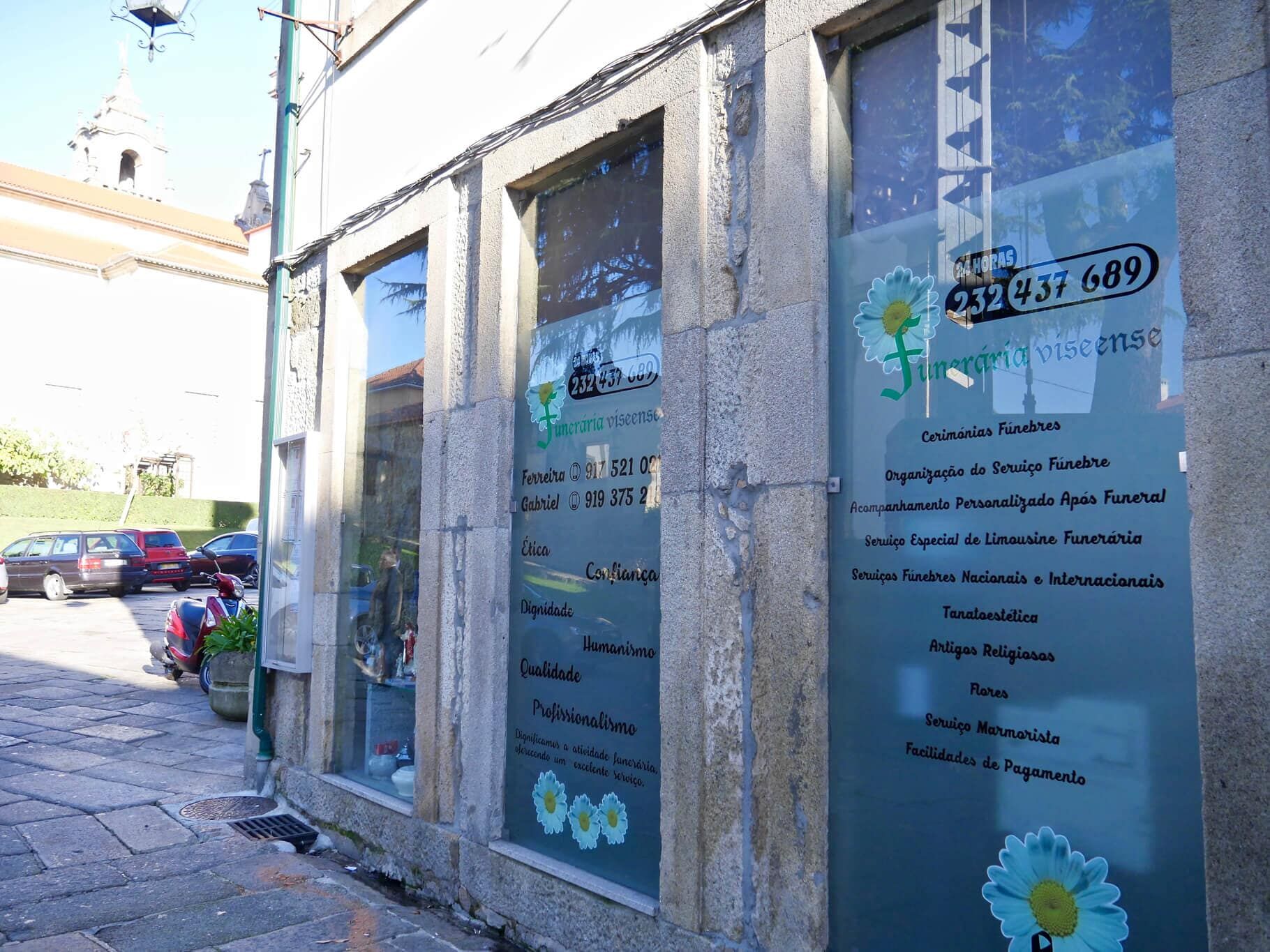 Fachada com janelas foscas, texto e logotipo de flores, ao lado de uma rua estreita com carros estacionados.