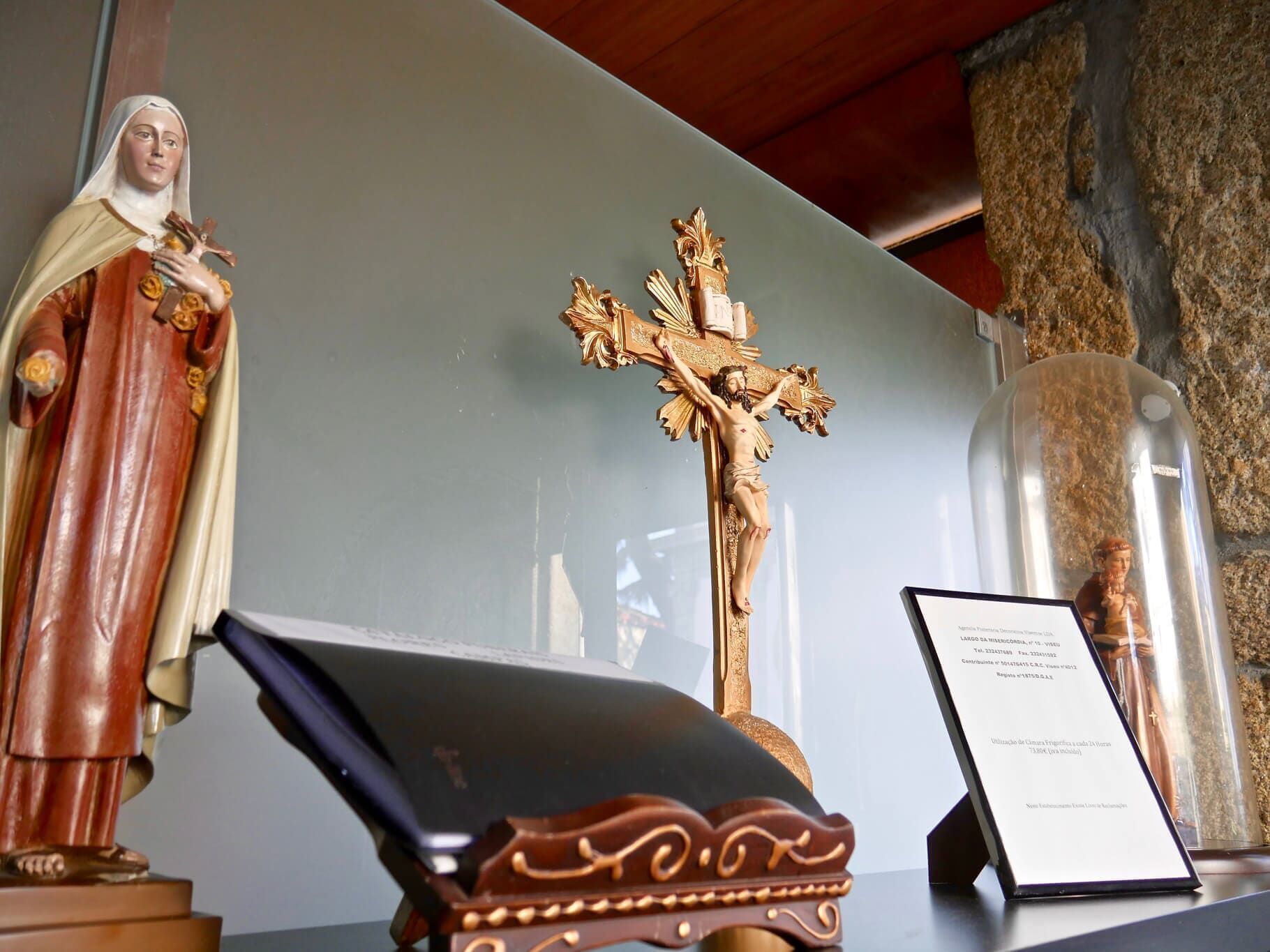 Estátuas de santo, crucifixo e livro ficam em uma prateleira com fundo de vidro.